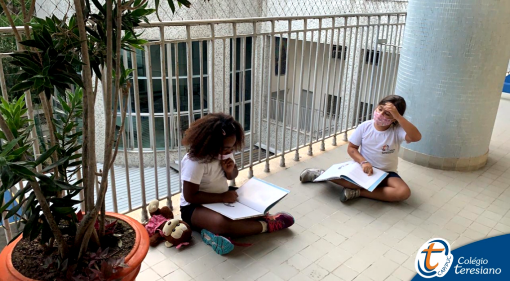 No mezanino do Colégio Teresiano, duas alunas estão sentadas com seus cadernos, estudando juntas. Ambas usam máscaras.