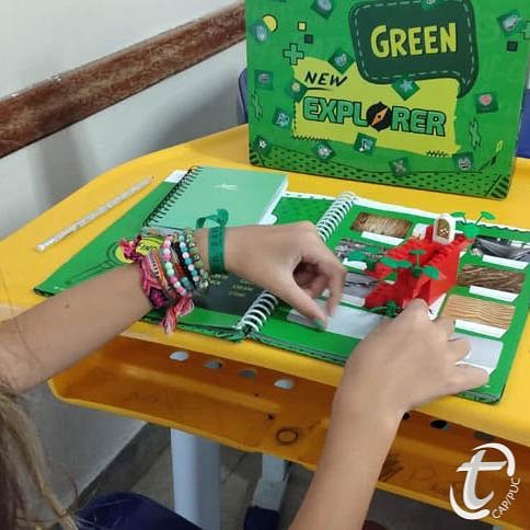 Em close na apostila verde, vemos uma aluna do Colégio Teresiano praticando colagens em uma atividade bilíngue. Acima da mesa, há uma caixa adesivada com os dizeres “Green – New Explorer”.