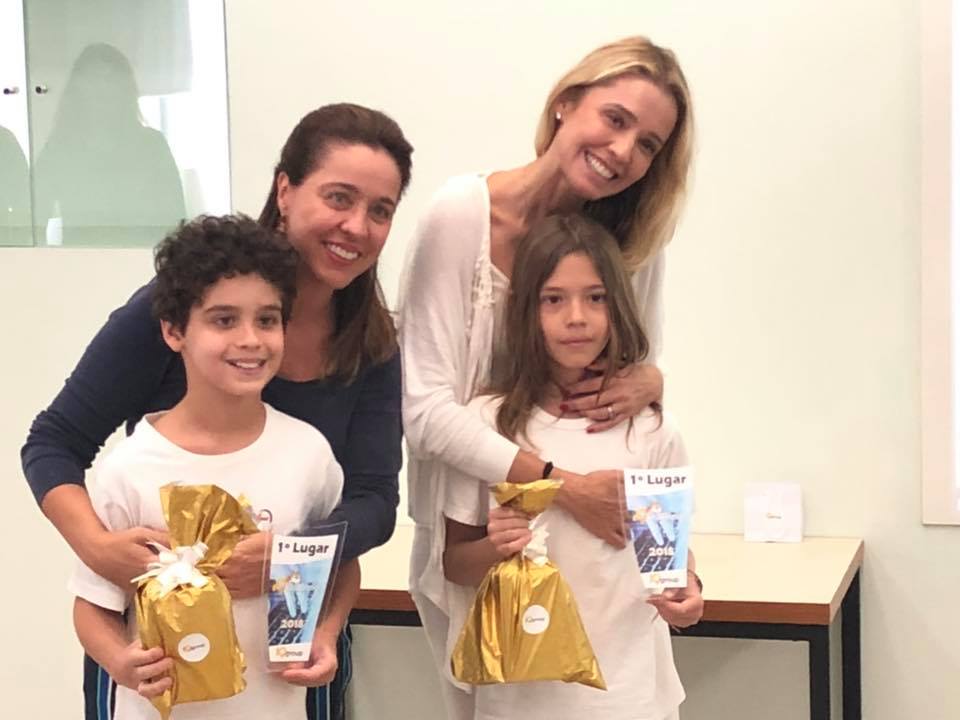 Foto de dois alunos, um menino e uma menina, acompanhados por suas respectivas mães ao receber um troféu de primeiro lugar.